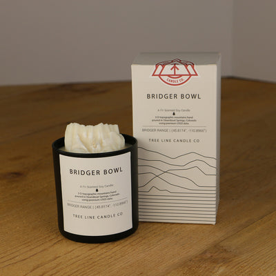 A white wax replica candle of Bridger Bowl next to a white box with red and black lettering.