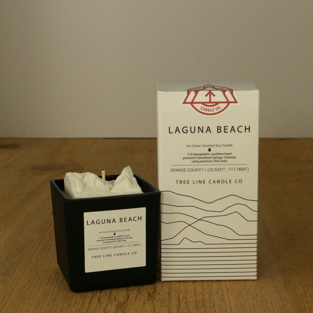 A white wax candle named Laguna Beach is next to a white box with red and black lettering.