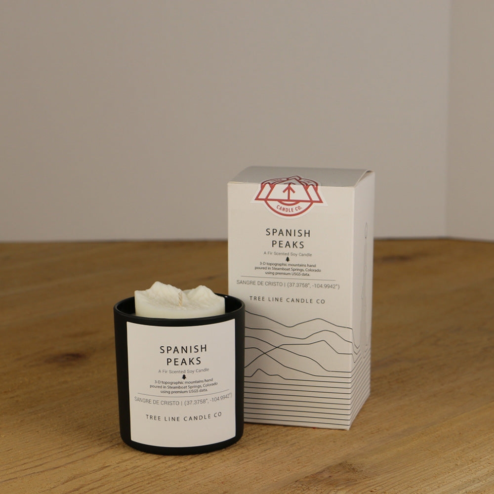 A white wax replica candle of Spanish Peaks summits next to a white box with red and black lettering.