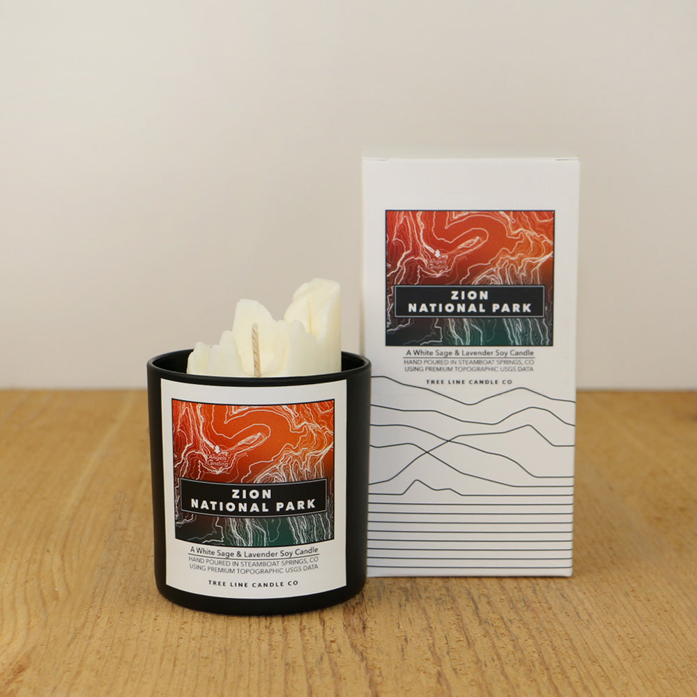 A white wax candle of Angels Landing with an orange, red, and green label is next to a white box with topography lines named Zion National Park by Tree Line Candle Co.