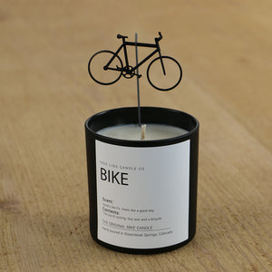 A white wax candle in a black container has a small bicycle placed over it.