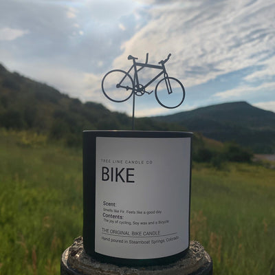 A black replica of a bicycle attachment is placed over a smooth wax candle in a black glass against a mountain background.