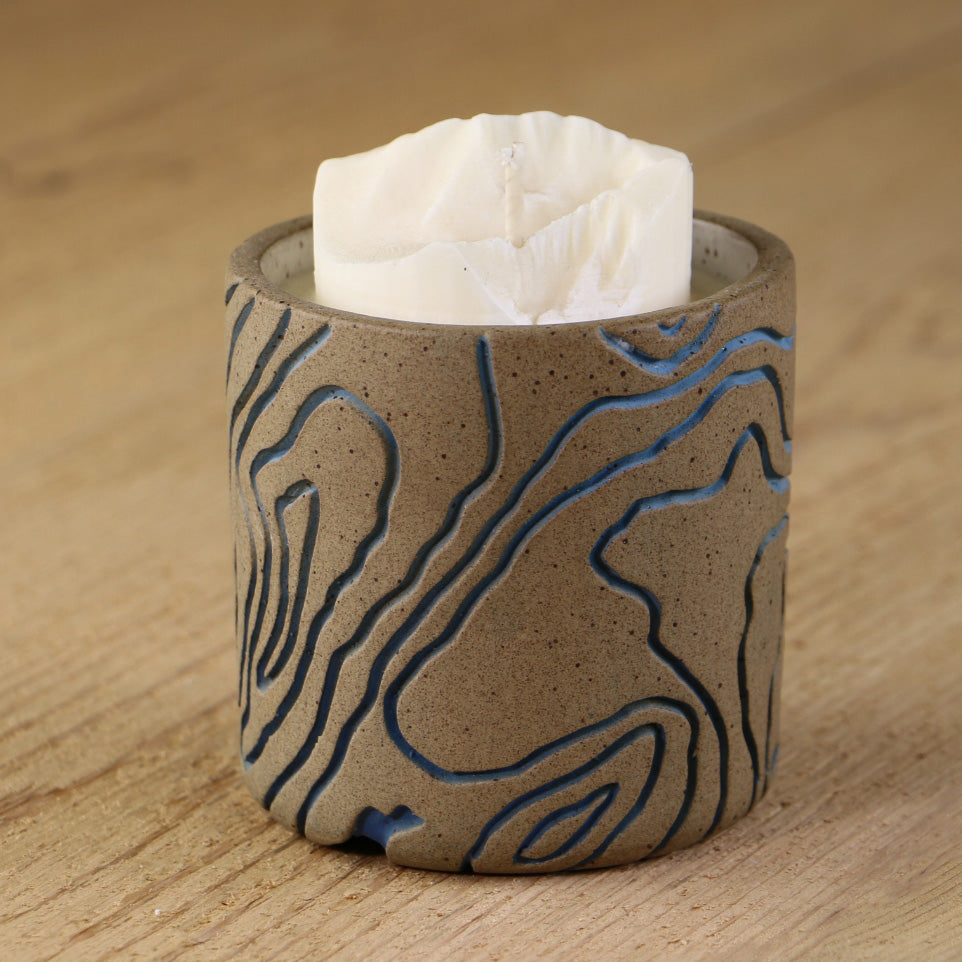 A white soy wax candle replica of Mount Evans in a decorative ceramic base inlaid with blue lines.