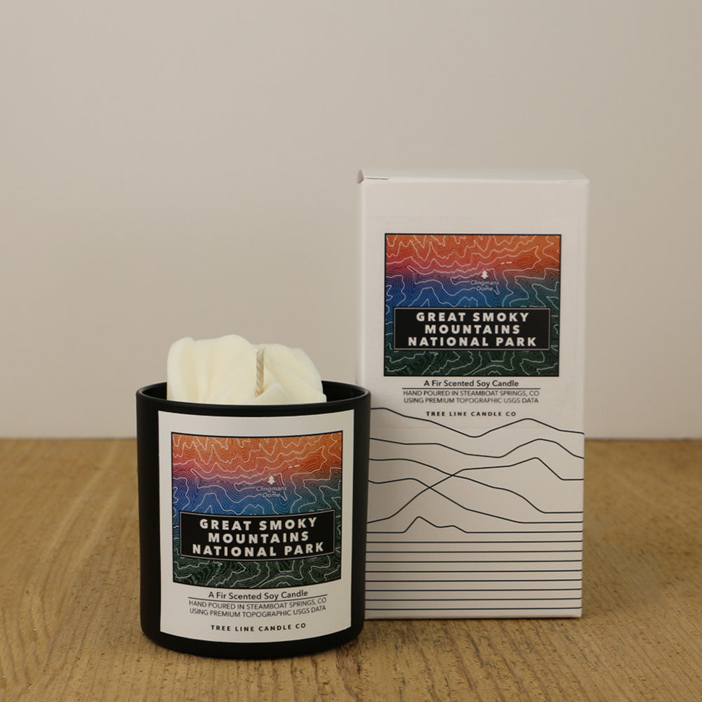 A white wax candle of Clingmans Dome with an orange, red, blue and green label is next to a white box with topography lines named Great Smoky Mountains National Park by Tree Line Candle Co.