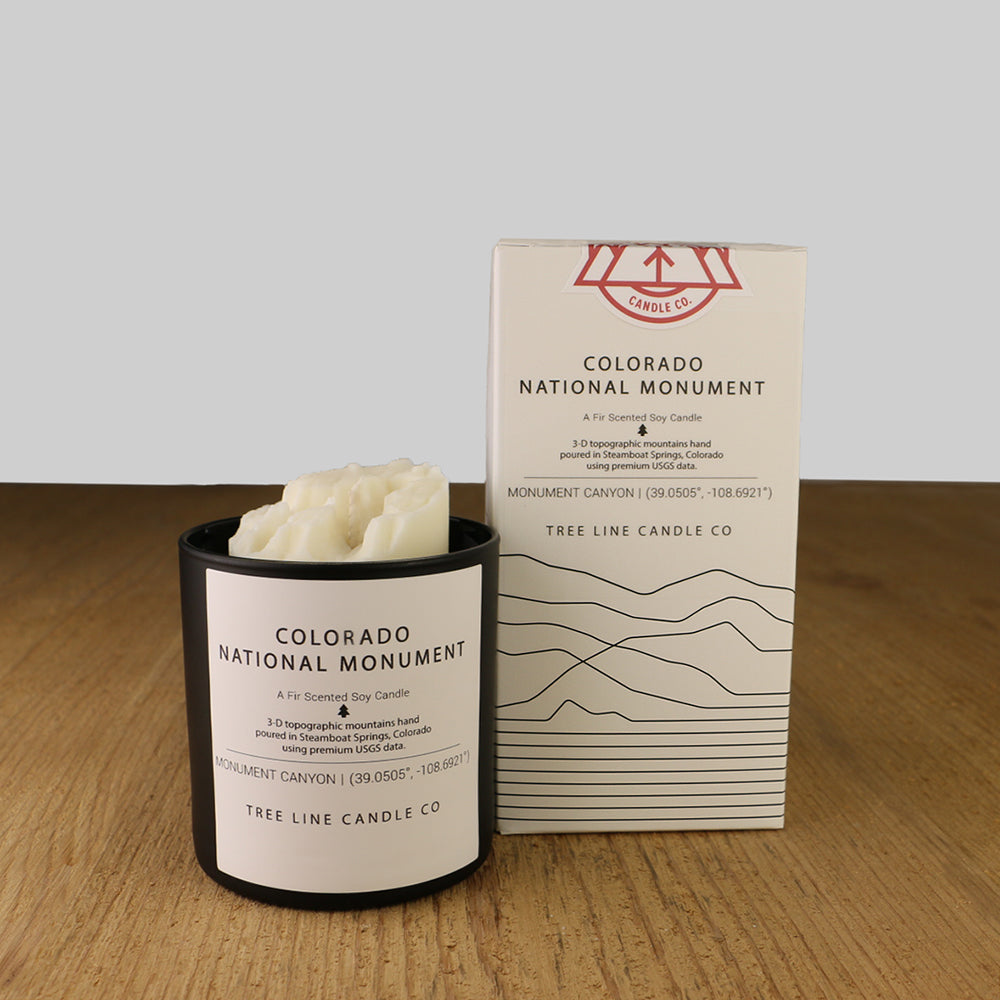 A white wax candle named Colorado National Monument is next to a white box with red and black lettering.