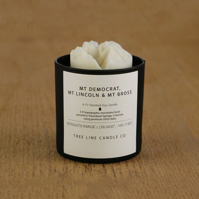 A white soy wax replica candle of Mt. Democrat, Mt. Lincoln & Mt Bross in a round, black glass.