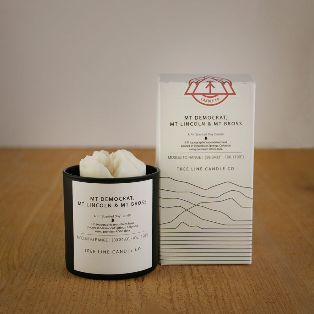 A white wax candle named  Mt. Democrat, Mt. Lincoln & Mt Bross is next to a white box with red and black lettering.