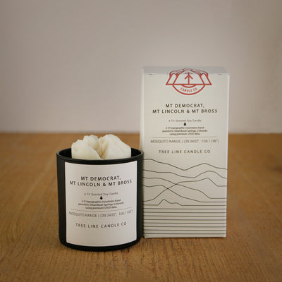 A white wax candle named  Mt. Democrat, Mt. Lincoln & Mt Bross is next to a white box with red and black lettering.