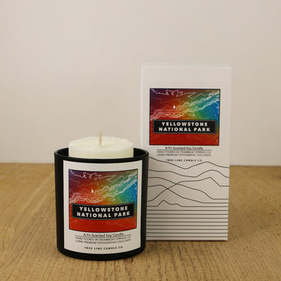 A white wax candle of Grand Canyon with an orange, blue and green label is next to a white box with topography lines named Yellowstone National Park by Tree Line Candle Co.