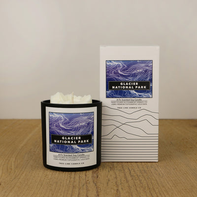 A white wax candle of Mount Grinnell	 with a purple and black label is next to a white box with topography lines named Zion National Park by Tree Line Candle Co.