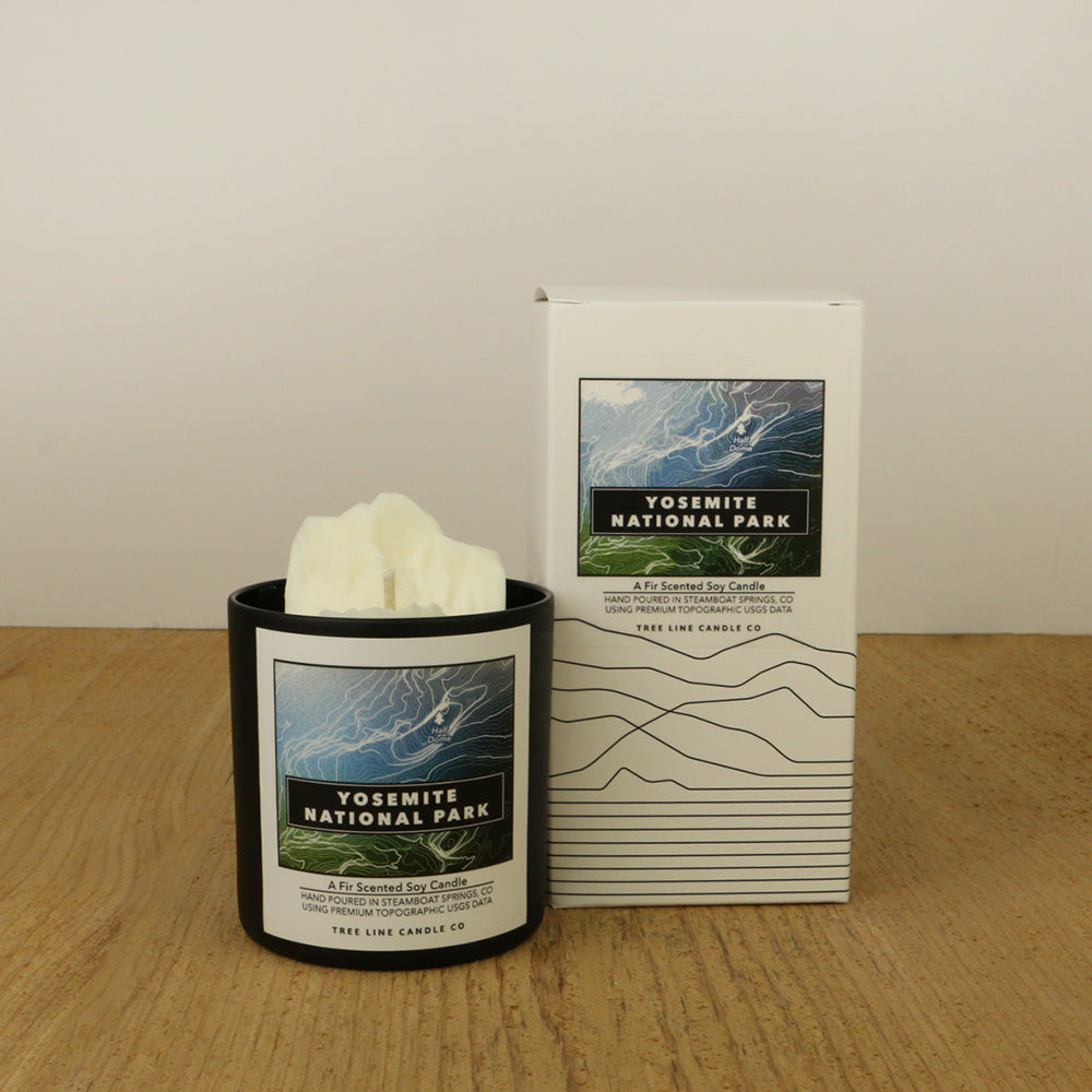 A white wax candle of Half Dome with a blue, black and green label is next to a white box with topography lines named Yosemite National Park by Tree Line Candle Co.