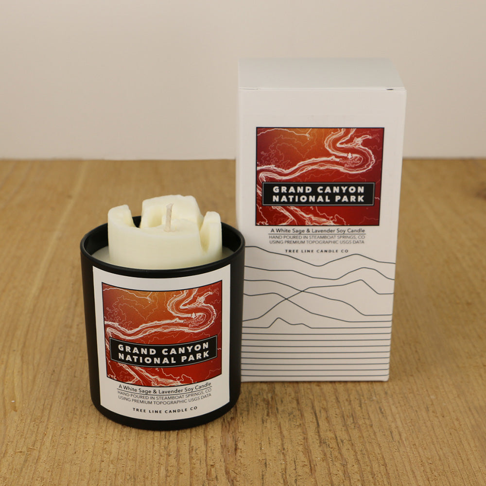 A white wax candle of Horseshoe Bend with a red and orange label is next to a white box with topography lines named Grand Canyon National Park by Tree Line Candle Co.