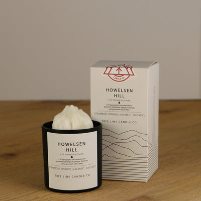 A white wax replica candle of Howelsen Hill summit next to a white box with red and black lettering.