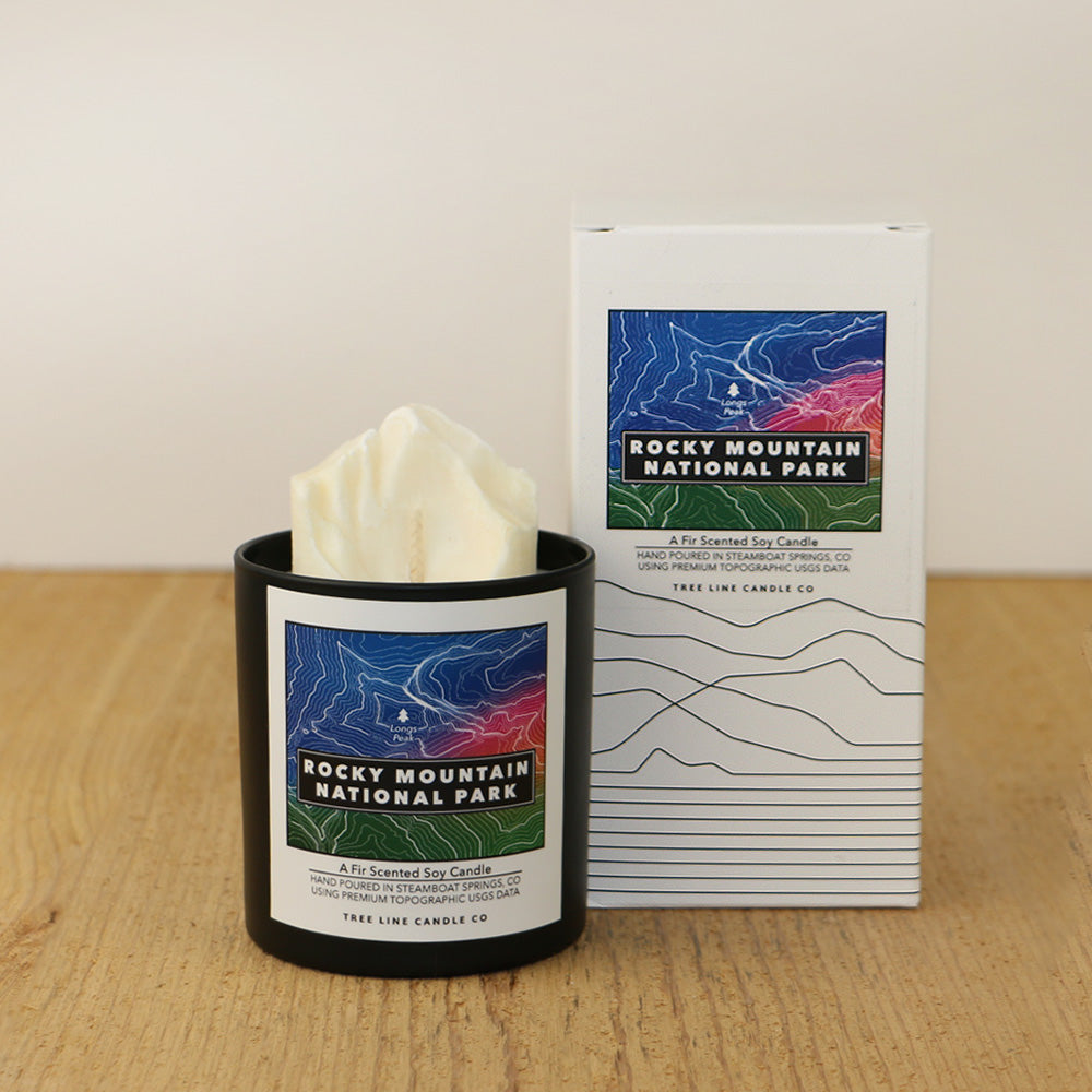 A white wax candle of Longs Peak named Rocky Mountain National Park with a blue, red and green label is next to a white box with topography lines by Tree Line Candle Co.