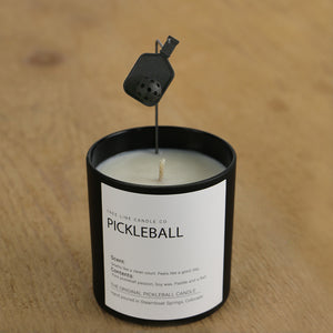 A white candle in a black glass container with a Pickleball paddle and ball attached over it.