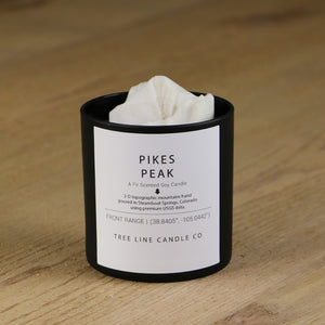 A white soy wax replica candle of Pikes Peak in a round, black glass.