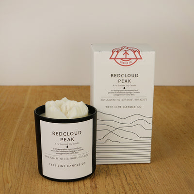 A white wax candle named Red Cloud Peak is next to a white box with red and black lettering.