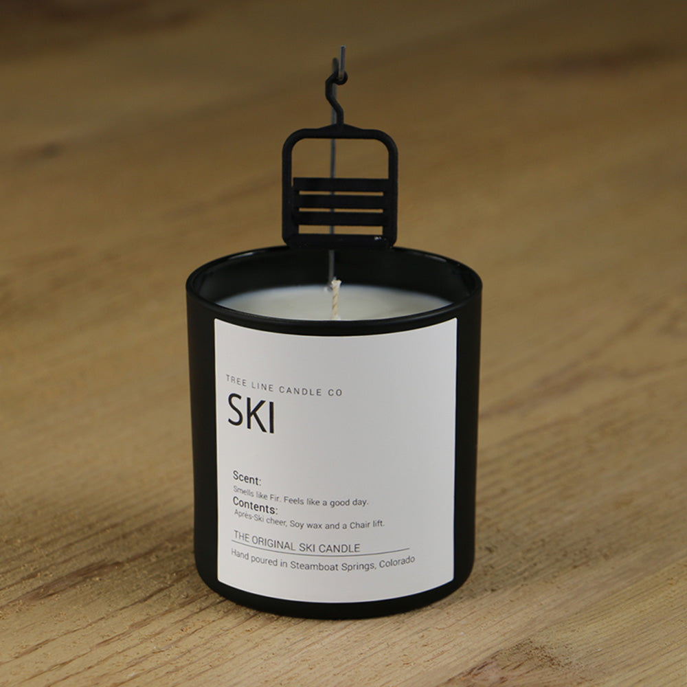 A white soy wax candle named Ski candle with a small black gondola chair hanging down in a round, black glass.