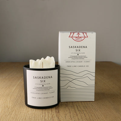 A white wax candle named Saskadena Six is next to a white box with red and black lettering.