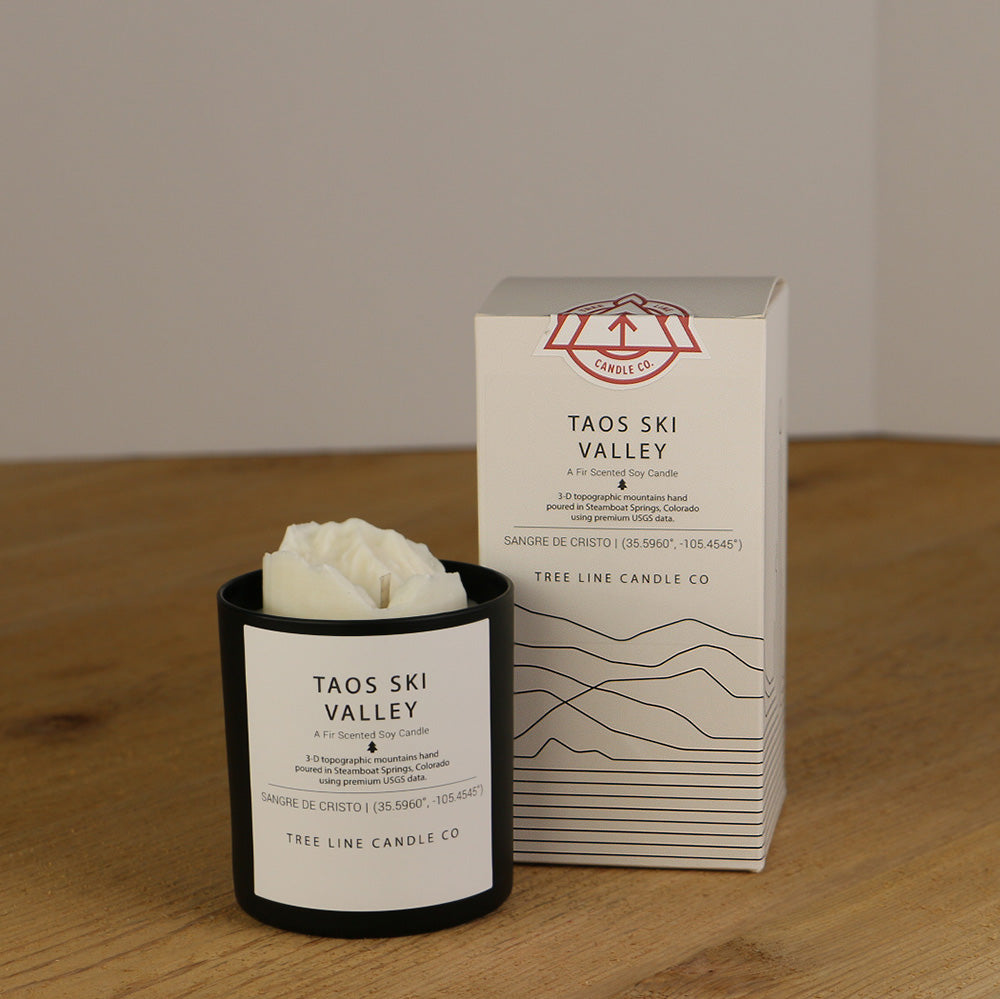A white wax replica candle of Taos Ski Valley next to a white box with red and black lettering.