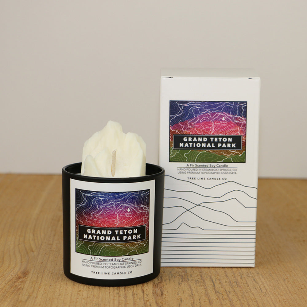 A white wax candle of Grand Teton with a blue, red and green label is next to a white box with topography lines named Grand Teton National Park by Tree Line Candle Co.